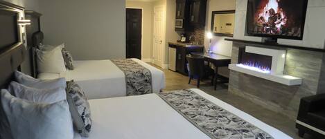 A hotel room with two beds, a desk, a television, and a fireplace.
