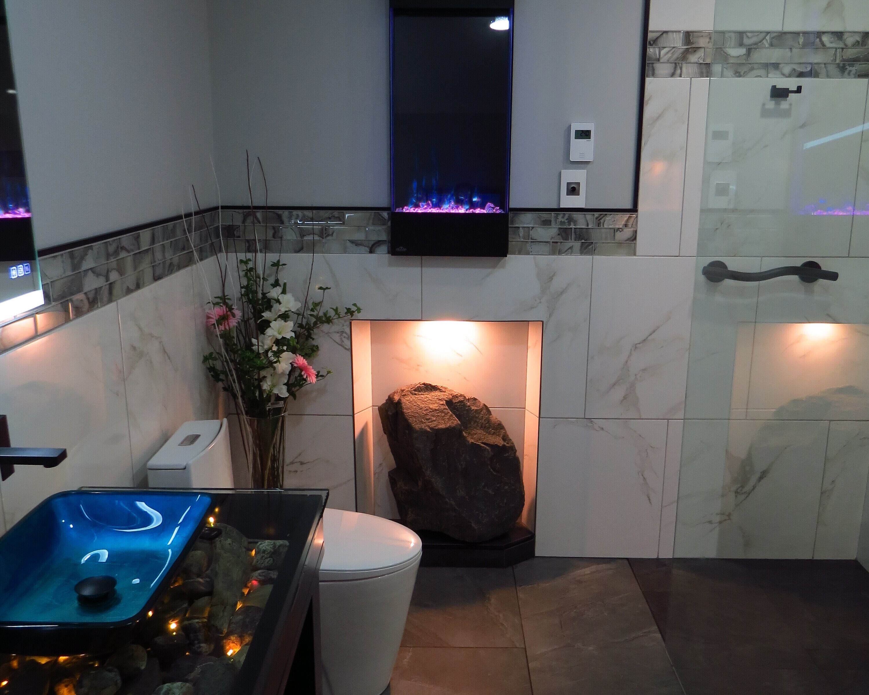 Un baño moderno con cabina de ducha de vidrio, inodoro blanco, una roca decorativa y un jarrón con flores.
