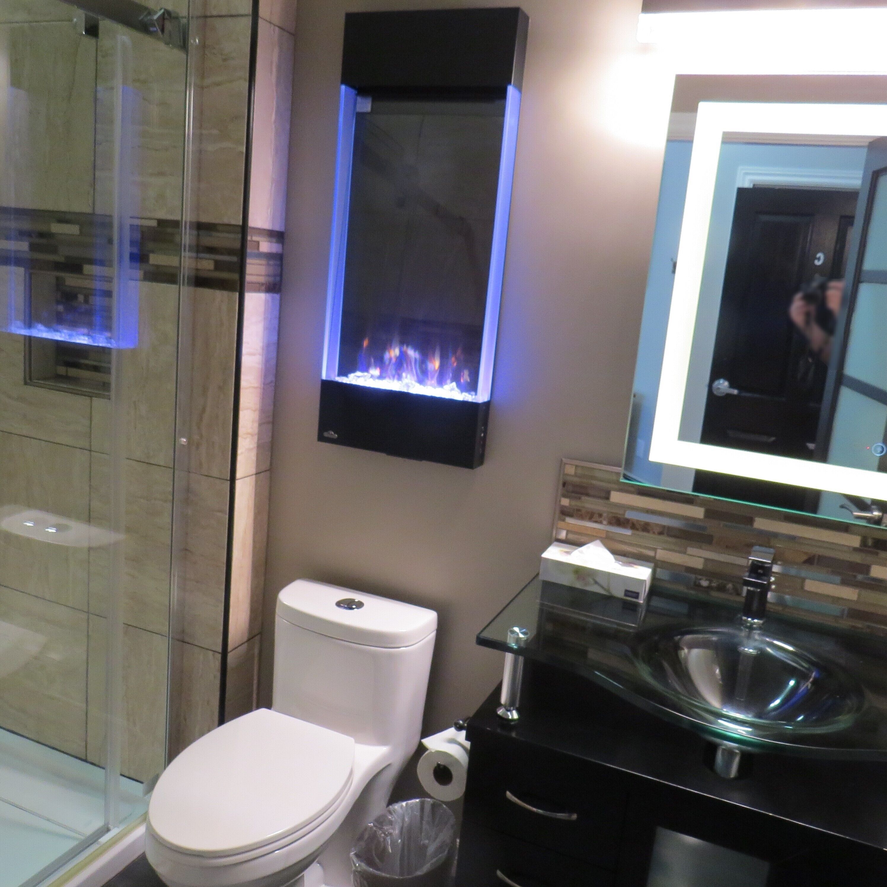 Une salle de bain moderne avec une douche à parois de verre, des toilettes, un lavabo en verre et un téléviseur fixé au mur qui diffuse une image de cheminée.