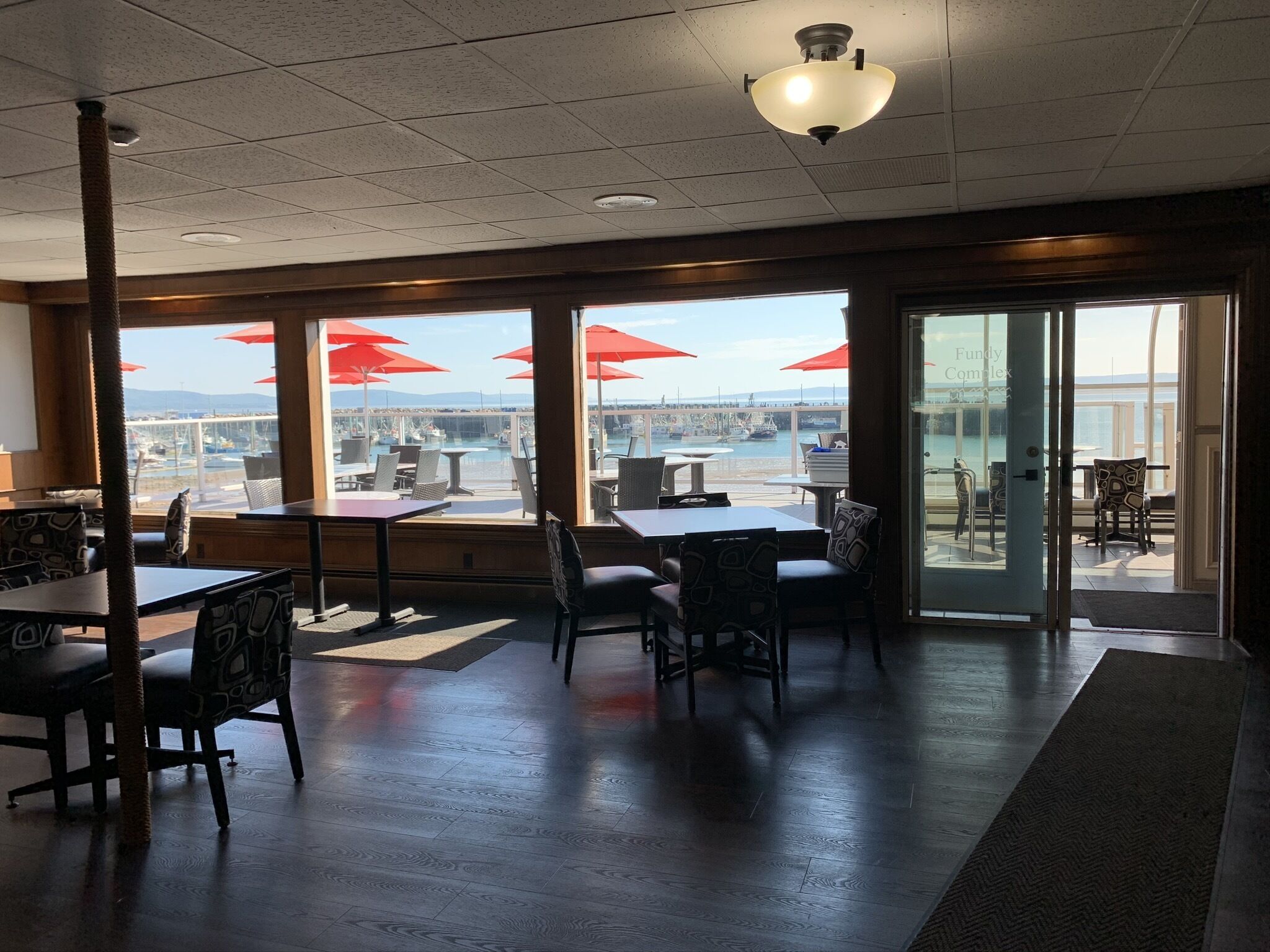 Un comedor interior con mesas y sillas, amplias ventanas con sombrillas rojas, y vista al puerto deportivo.
