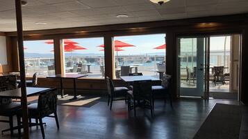 Un comedor interior con mesas y sillas, amplias ventanas con sombrillas rojas, y vista al puerto deportivo.