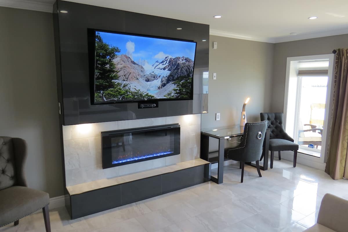 A modern living room with a flat-screen TV mounted above a fireplace, a dining area with a table and chairs, and a large window with curtains.