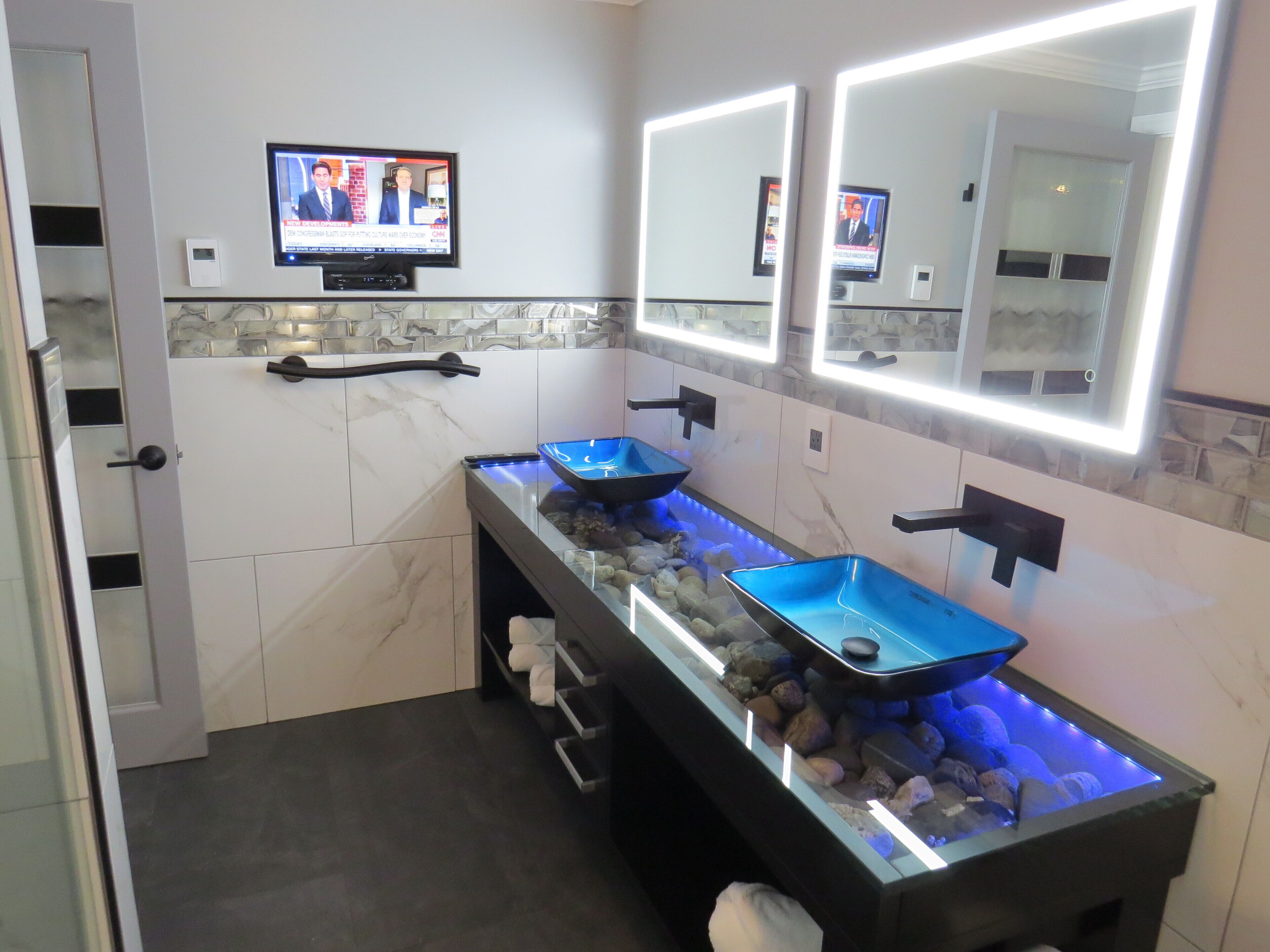 A modern bathroom with a glass sink, marble tiles, and a TV mounted above the sink.