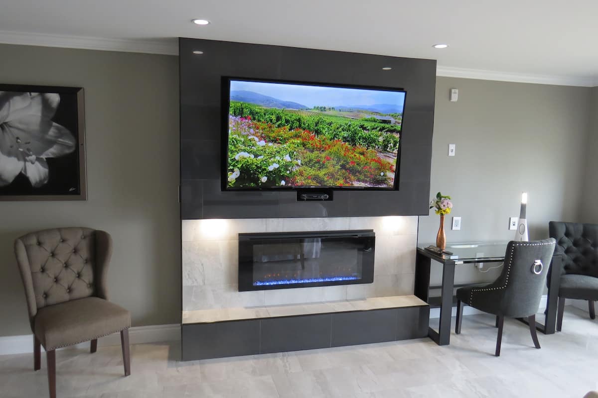 A modern living room with a flat-screen TV mounted above a fireplace, two upholstered chairs, and a table with a vase.