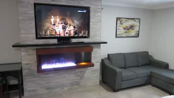 Een woonkamer met een flatscreen televisie aan een stenen muur, een open haard met vlammen, en een grijze bank.