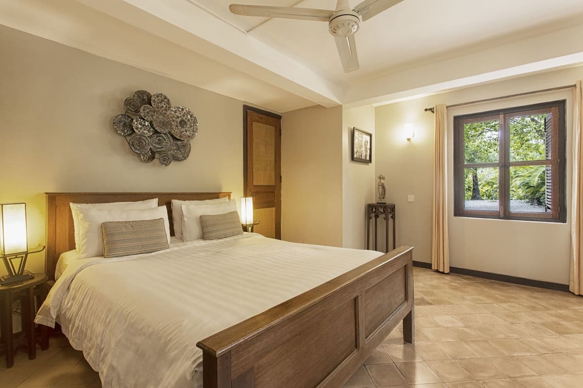 A bedroom with a wooden bed, a ceiling fan, a decorative wall piece, a window with curtains, and a nightstand with a lamp.