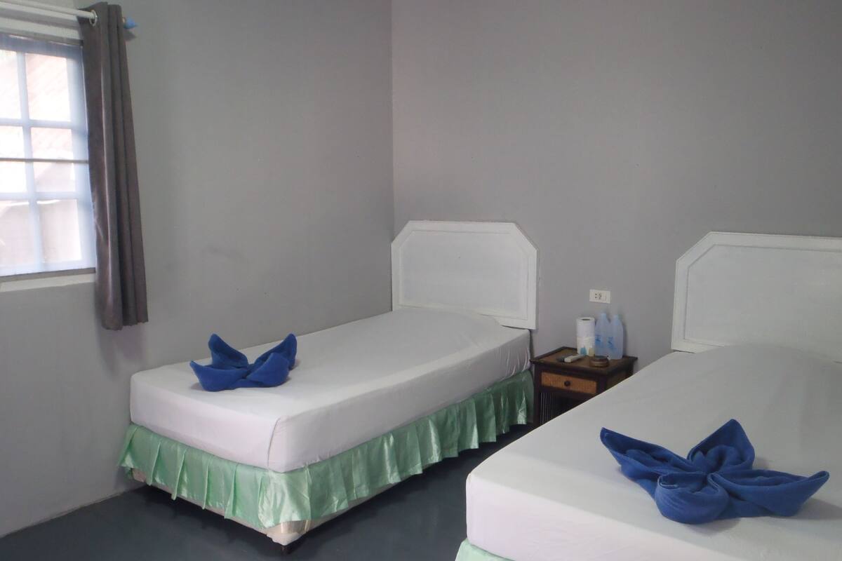 Ein Zimmer mit zwei Betten, jeweils mit weißer Matratze und grünem Betttuch. Ein Nachttisch mit Fernbedienung und Toilettenartikeln ist vorhanden.