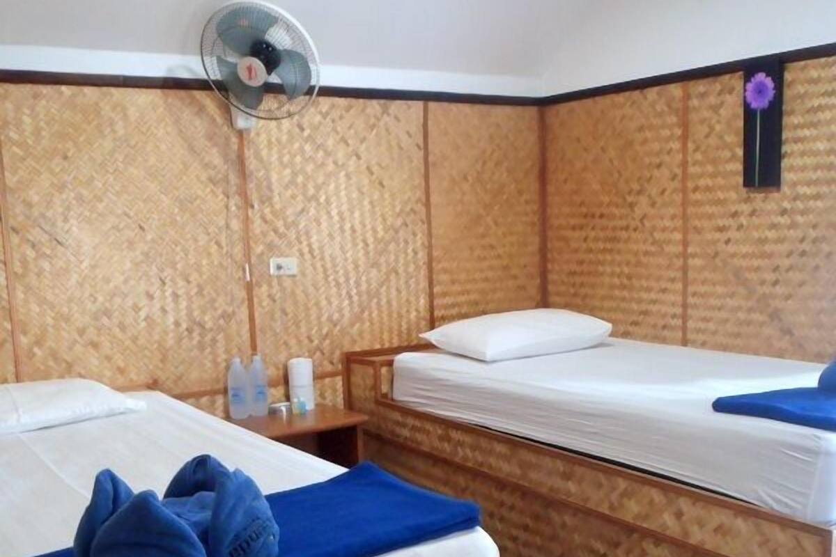 Ein Zimmer mit zwei Betten, einem Wandventilator und einem kleinen Tisch mit Pflegeartikeln.