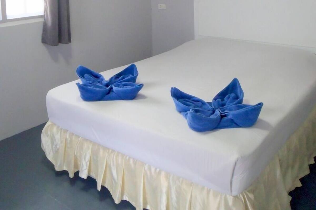 Ein Bett mit weißem Kopfteil und einem cremefarbenen Bettvorhang. Auf dem Bett liegen zwei blaue, gefaltete Handtücher.