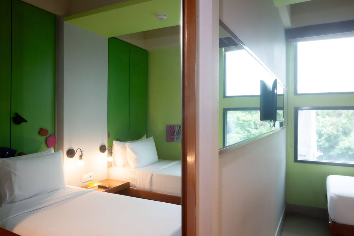 A room with two beds, a green accent wall, a large window, and a mounted television.