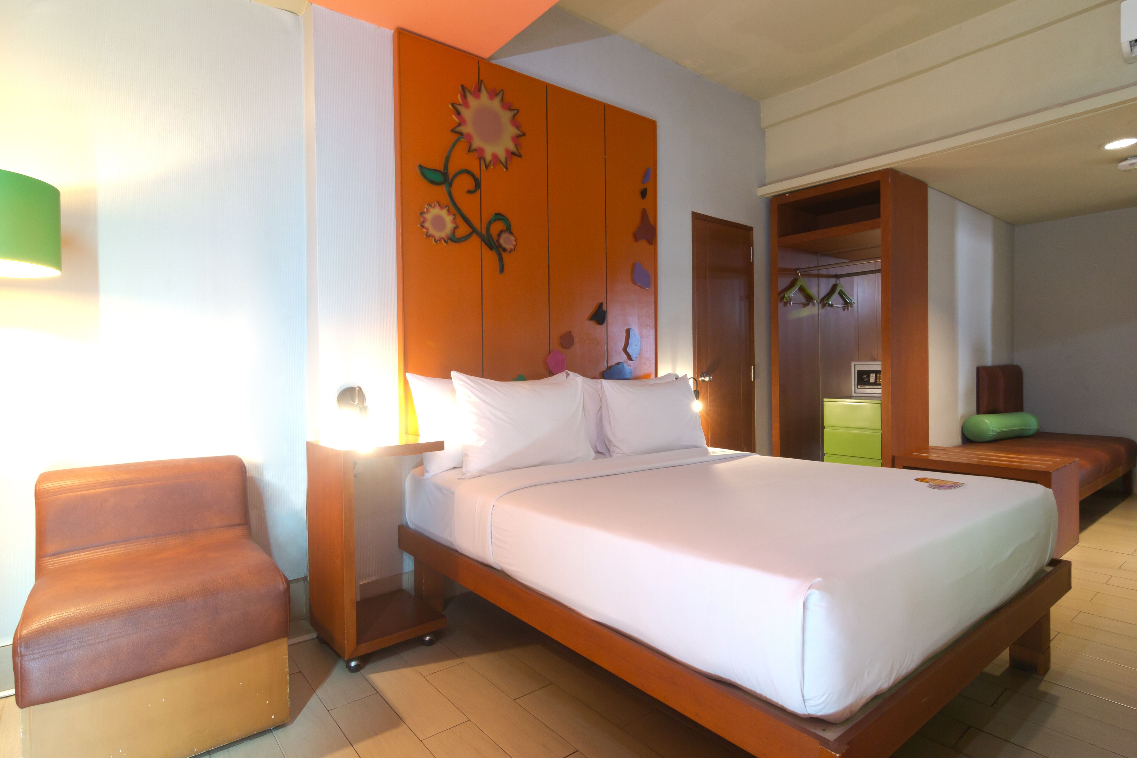 A neatly made bed with white linens, a wooden headboard, and a wall with a floral design. There is a wooden bench and a nightstand with a lamp.