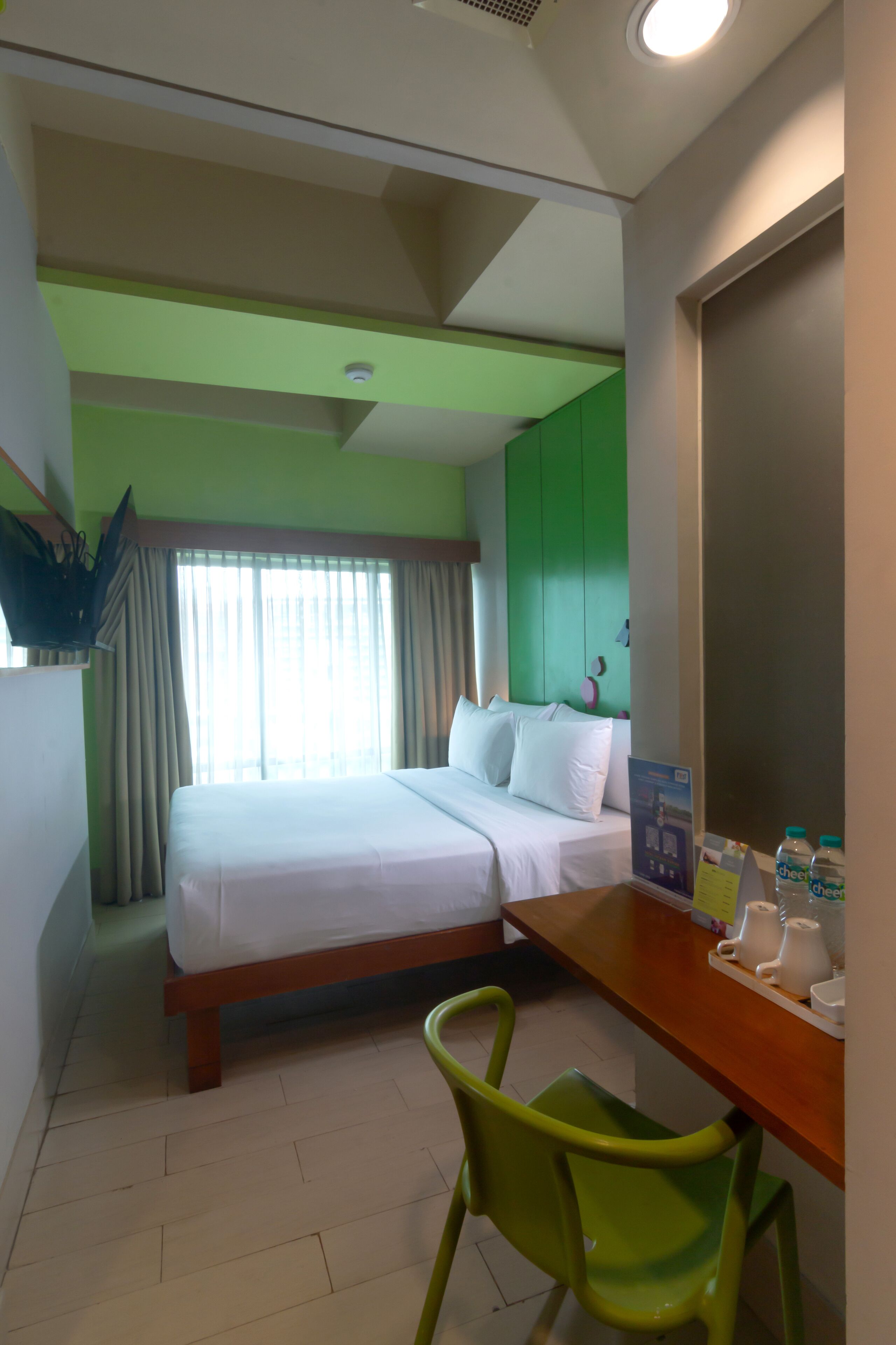 A hotel room with a bed, a desk, a chair, a window with curtains, and a wall-mounted TV.