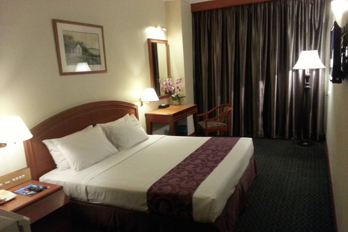 Quarto de hotel com uma cama grande, uma escrivaninha com luminária, uma cadeira, um espelho e uma janela com cortinas.