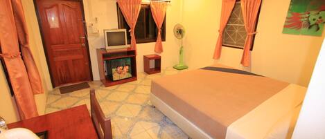Una habitación de hotel con una cama, televisión, una mesita, un ventilador y una puerta.