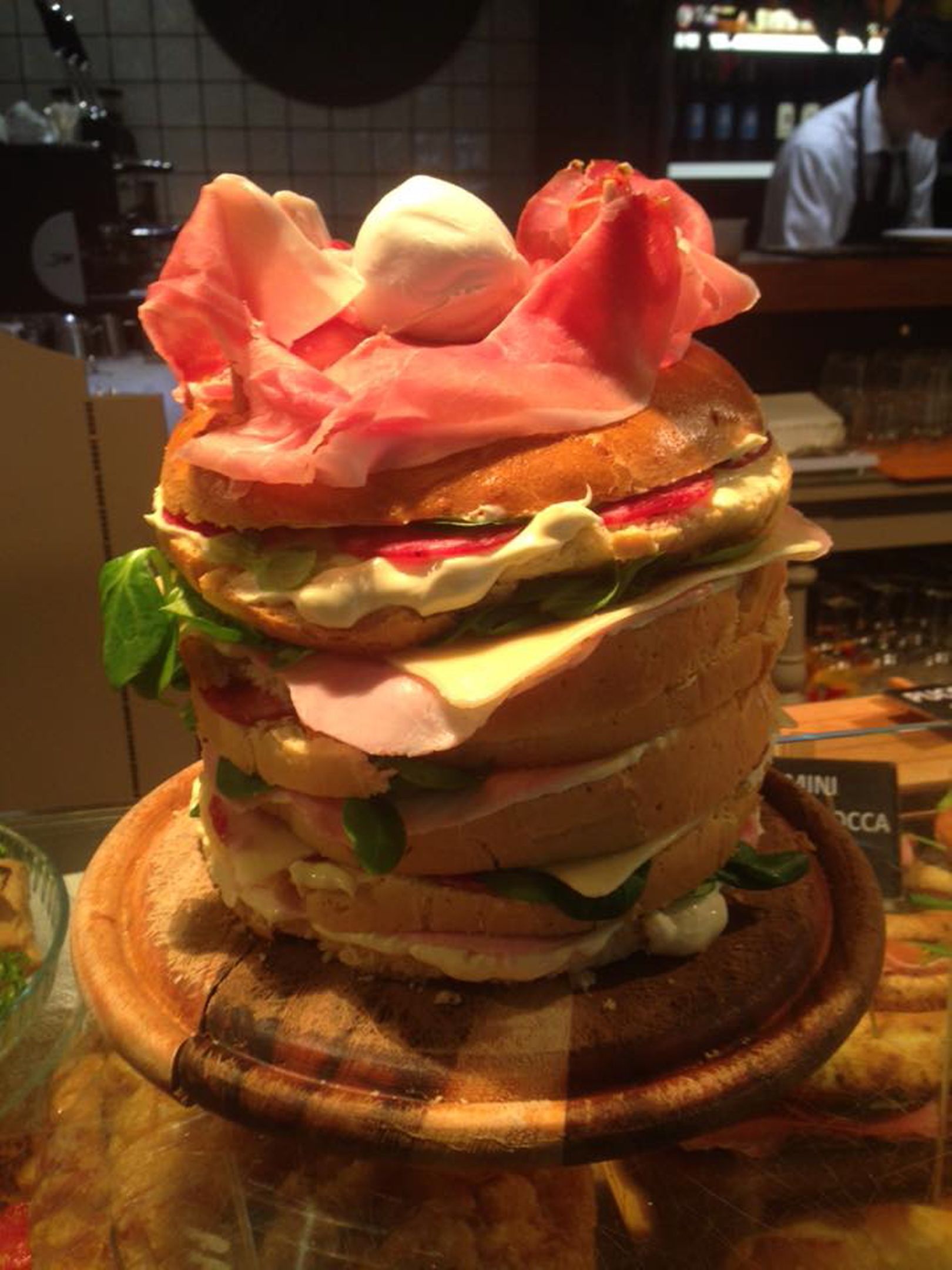 Un sándwich con varias capas de ingredientes, entre ellos lechuga, queso y carne, sobre una tabla de madera.