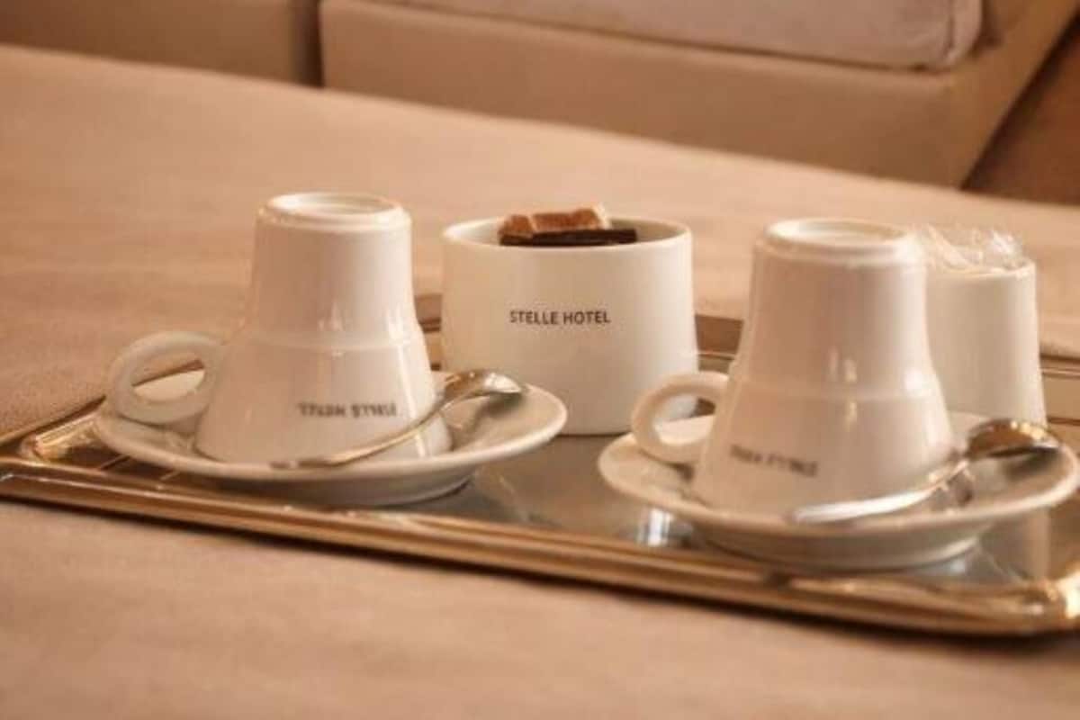 A hotel room with two beds, a tray with cups and a container labeled 'STELLE HOTEL,' and a bedside table with a lamp.