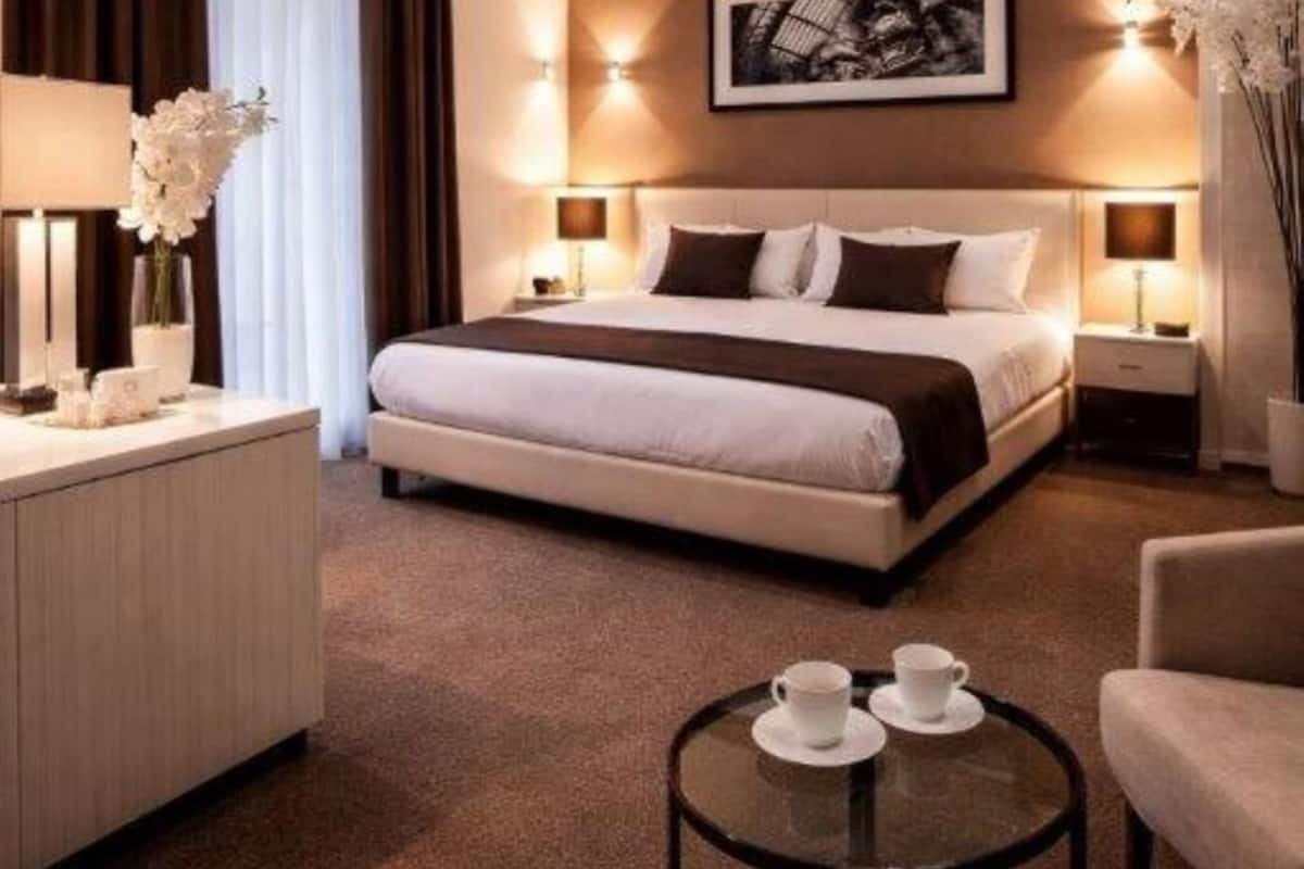 A modern hotel room with a large bed, two bedside tables with lamps, a desk, and a small round table with cups.