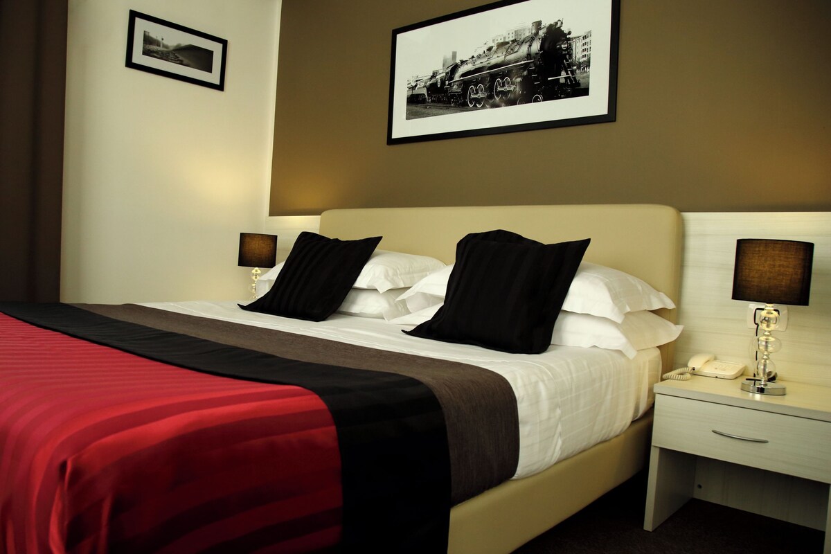 A hotel room with a bed, two bedside tables, a nightstand, and a lamp.