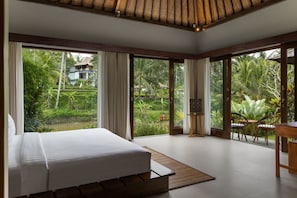 A bedroom with a large bed, a dining area with chairs, and a view of a lush garden. - Suara Air Resort Ubud (Ubud)