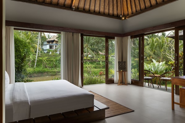 A bedroom with a large bed, a dining area with chairs, and a view of a lush garden. - Suara Air Resort Ubud (Ubud)