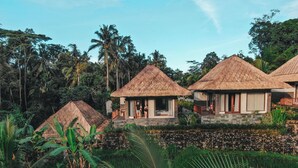 A tropical resort with thatched-roof huts, lush greenery, and a stone wall. - Suara Air Resort Ubud (Ubud)