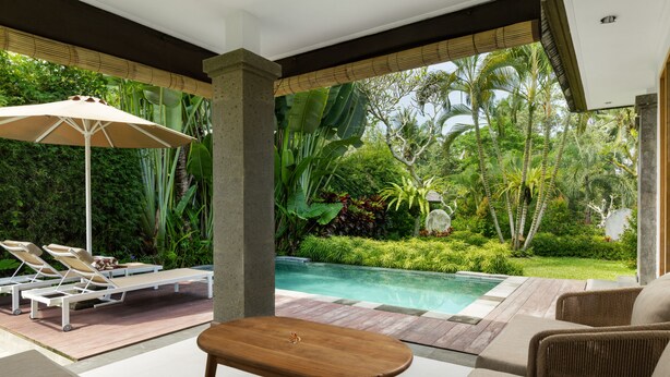 Zona junto a la piscina con sillones de descanso, una mesa de madera y vistas a una alberca rodeada de exuberante vegetación.