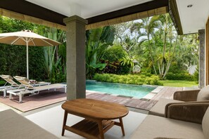 Zona junto a la piscina con sillones de descanso, una mesa de madera y vistas a una alberca rodeada de exuberante vegetación.