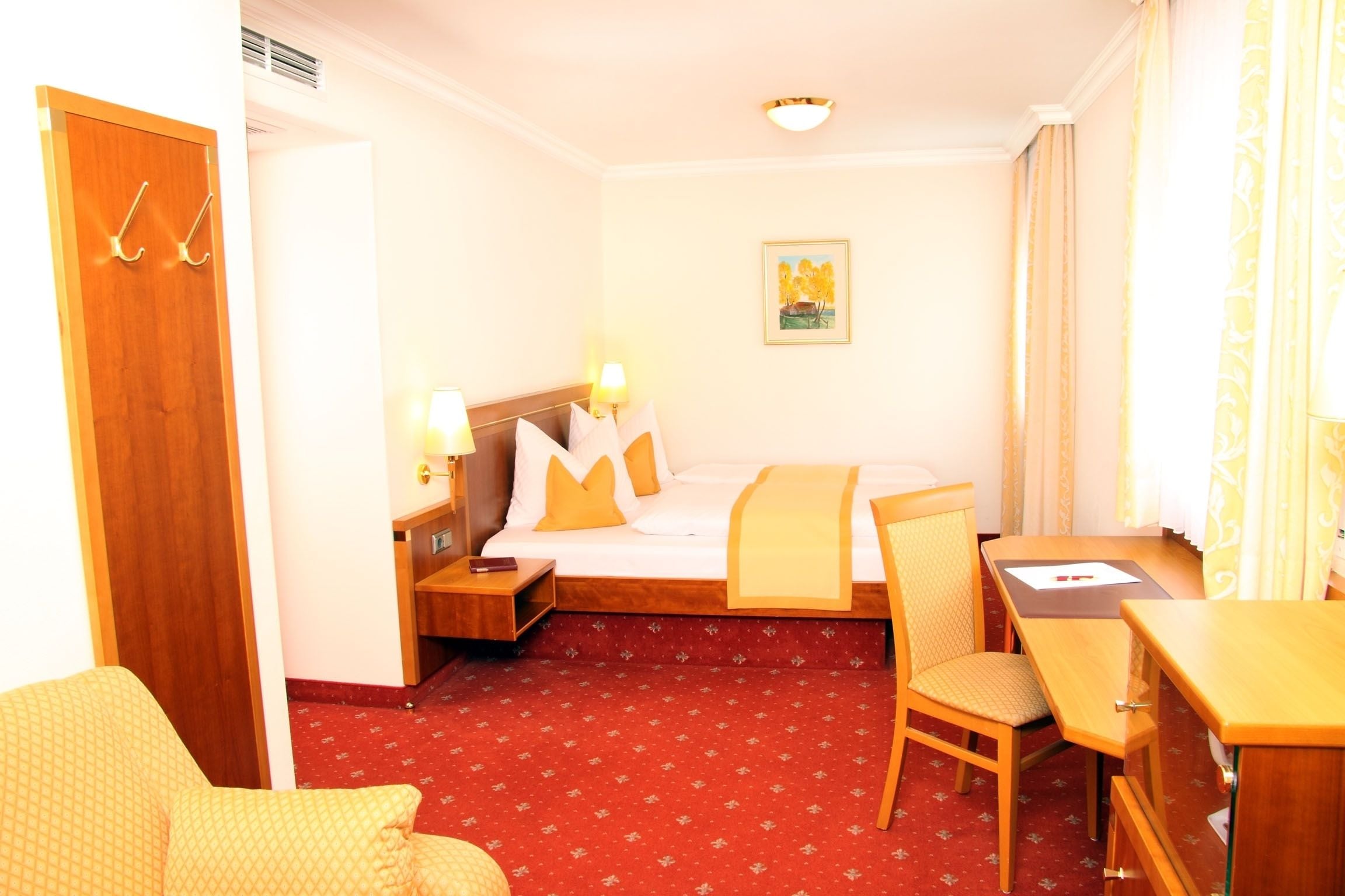 Habitación de hotel con cama, mesita de noche, lámpara, silla, escritorio y piso cubierto de alfombra roja.