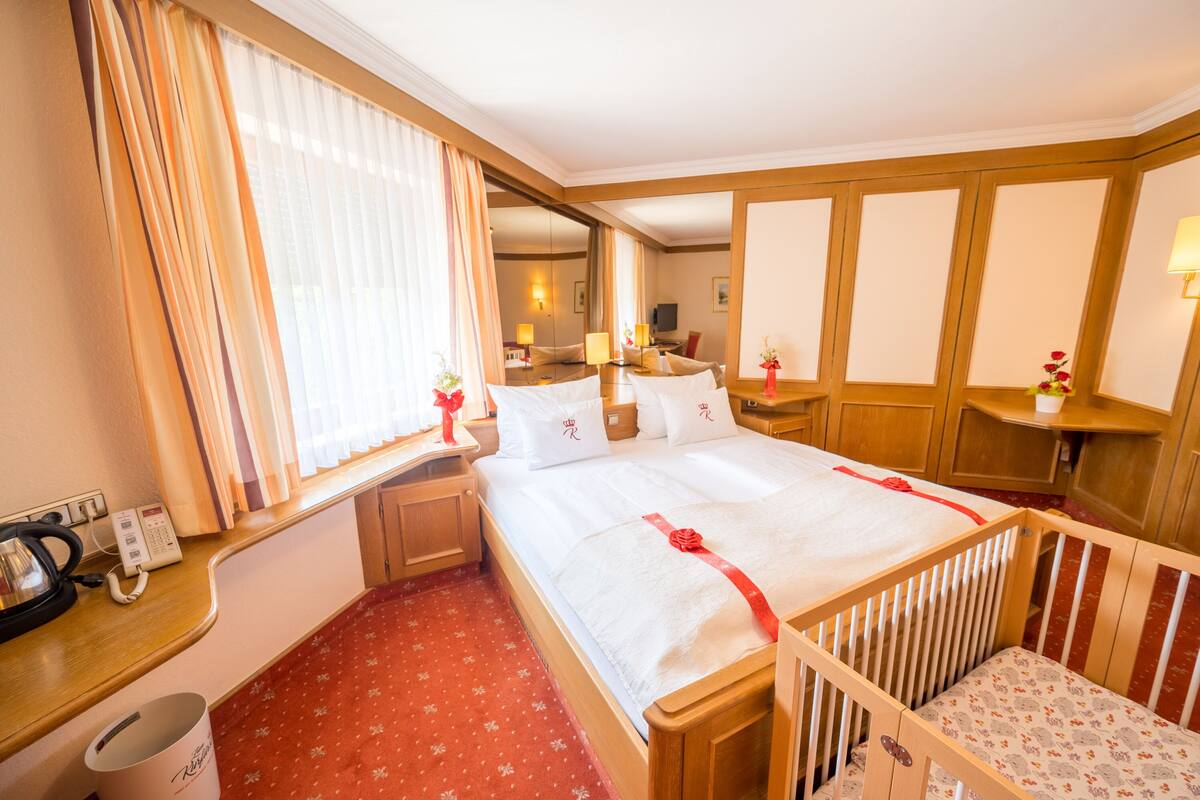 Une chambre d’hôtel avec un grand lit, un berceau en bois, un bureau avec un téléphone et une fenêtre avec des rideaux.