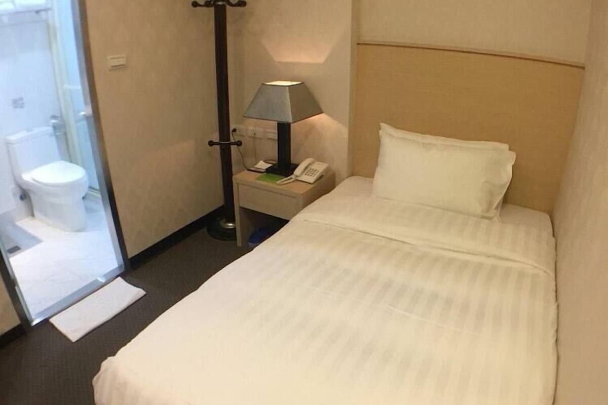 A hotel room with a bed, a nightstand, a lamp, a coat rack, and a bathroom visible through an open door.