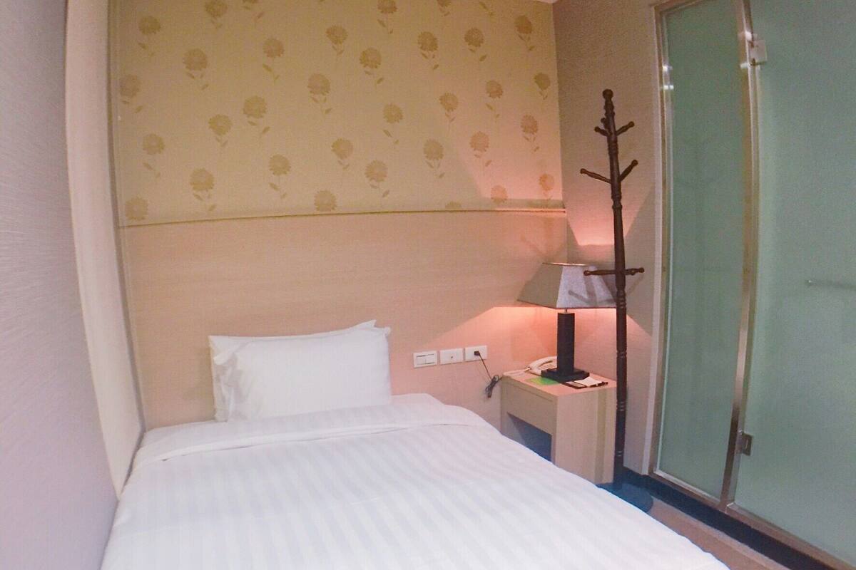 A single bed in a hotel room with a plain headboard, a bedside table with a lamp, and a door to a balcony.