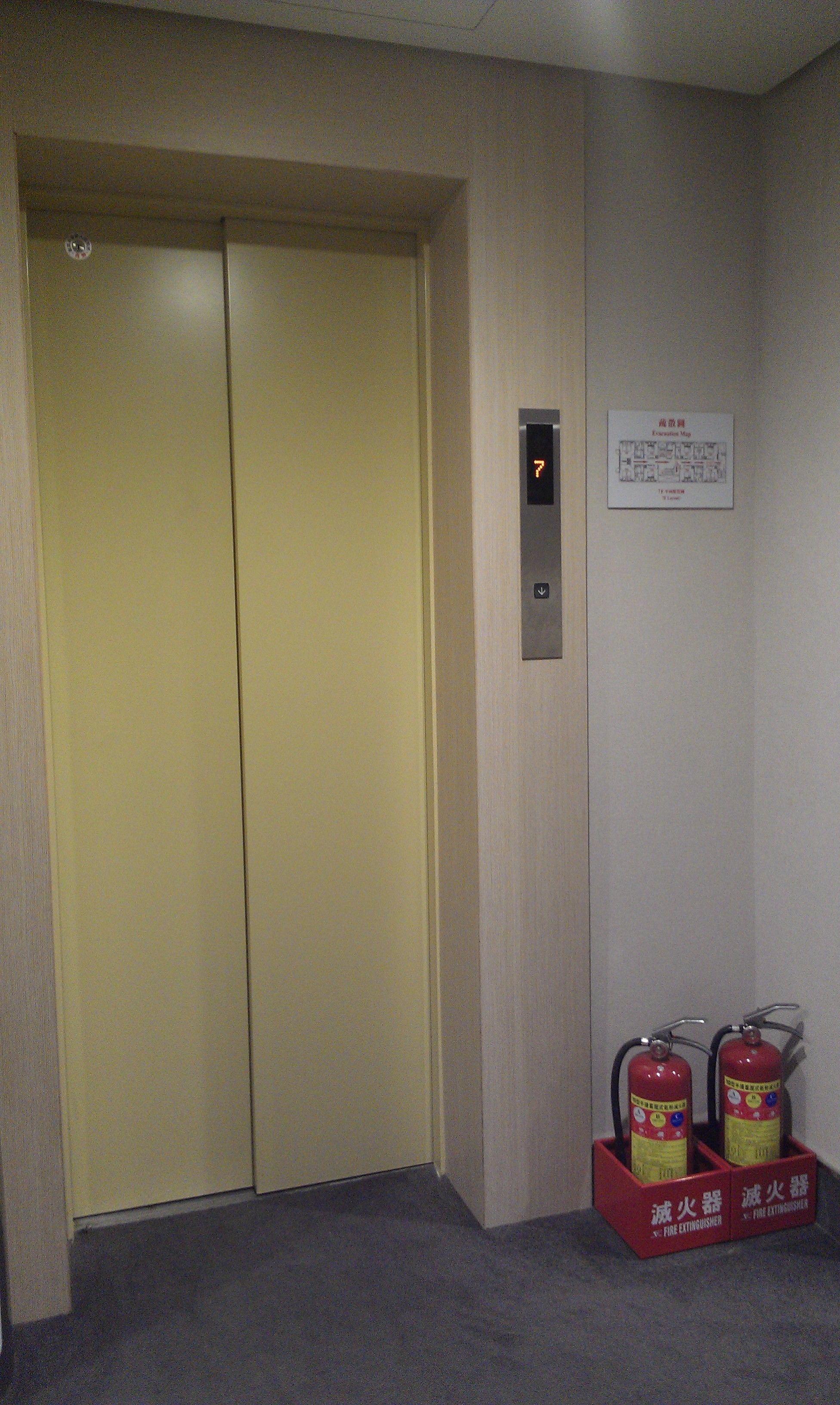 A hotel elevator with a yellow door, a fire extinguisher mounted on the wall, and an emergency exit sign.