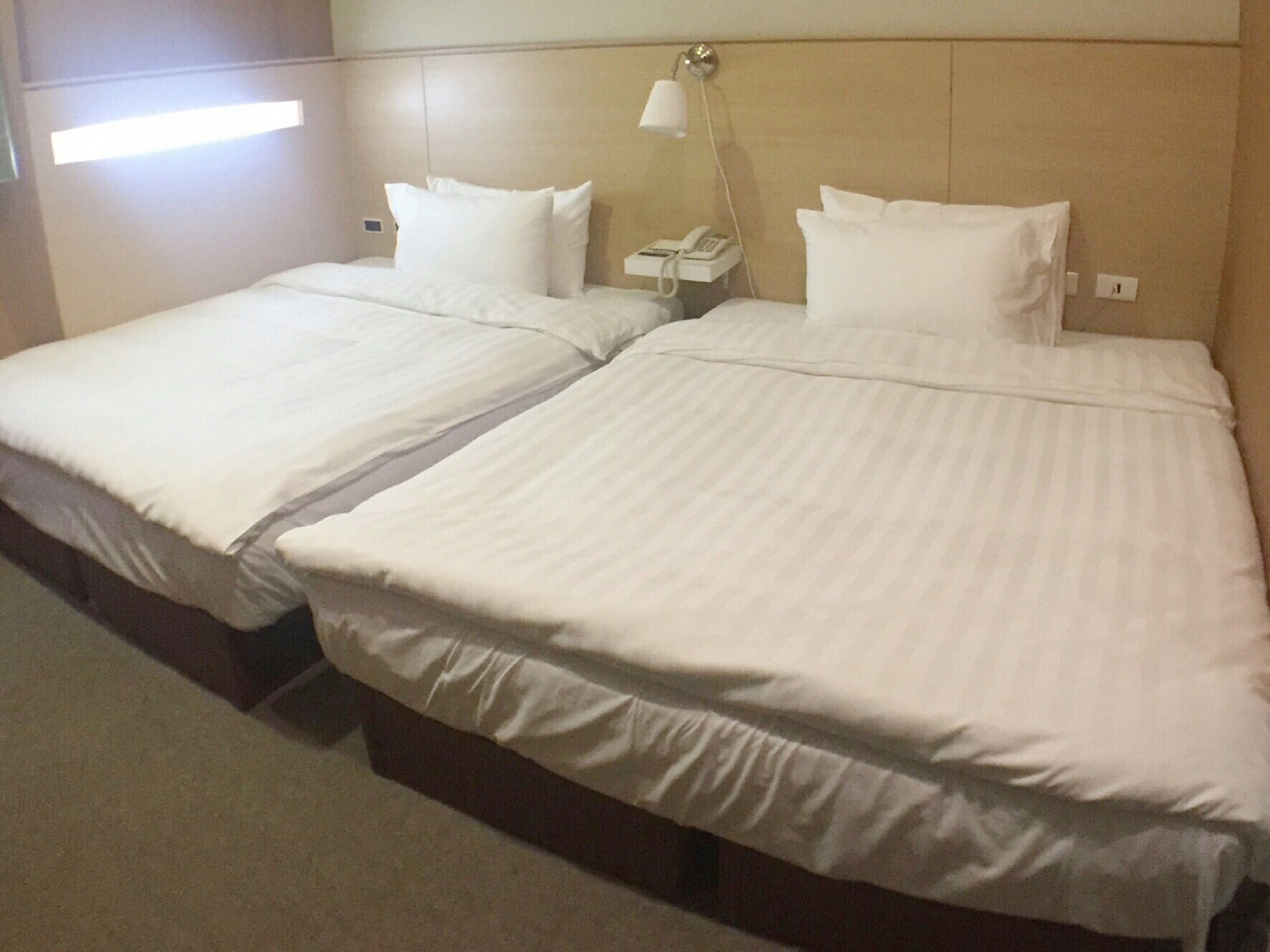 A hotel room with two single beds, each with white linens and pillows. There is a bedside table with a lamp and a phone.
