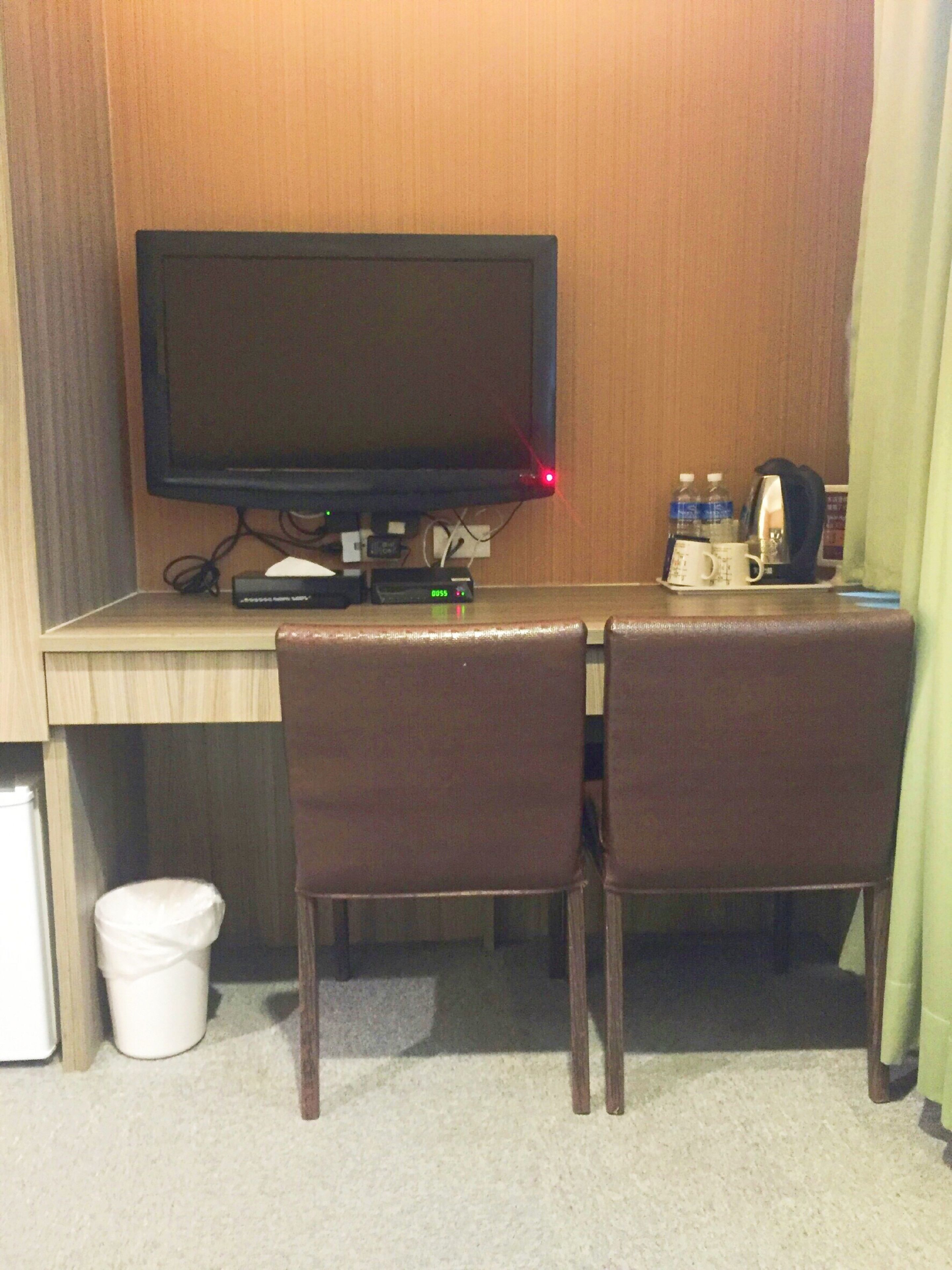 A hotel room with a desk, two chairs, a television, a coffee maker, and a trash bin.