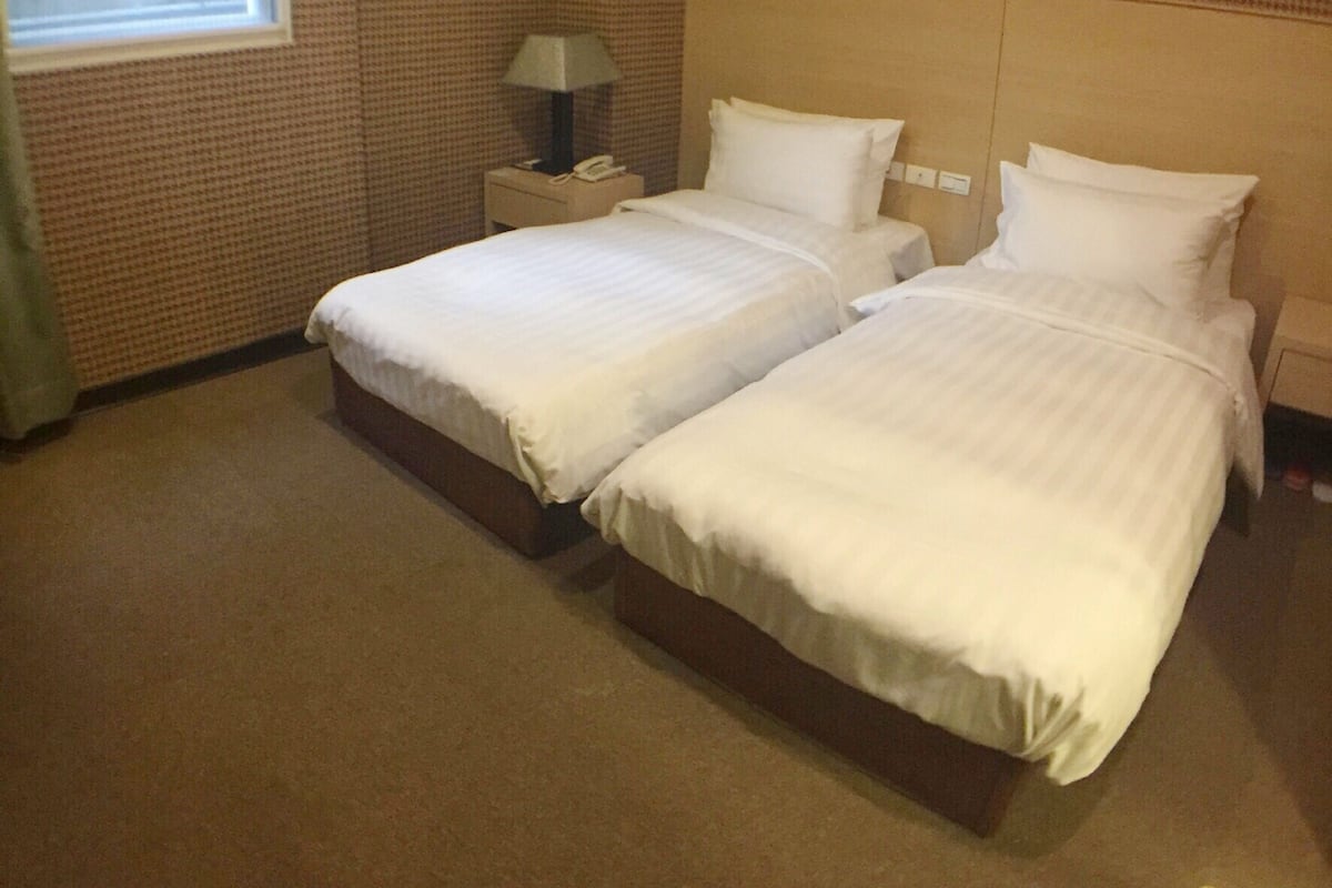 A hotel room with two beds, a nightstand with a lamp, and a window with blinds.