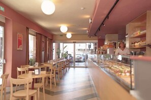 Um restaurante com mesas, cadeiras, um balcão com comida à mostra e vista para a rua.