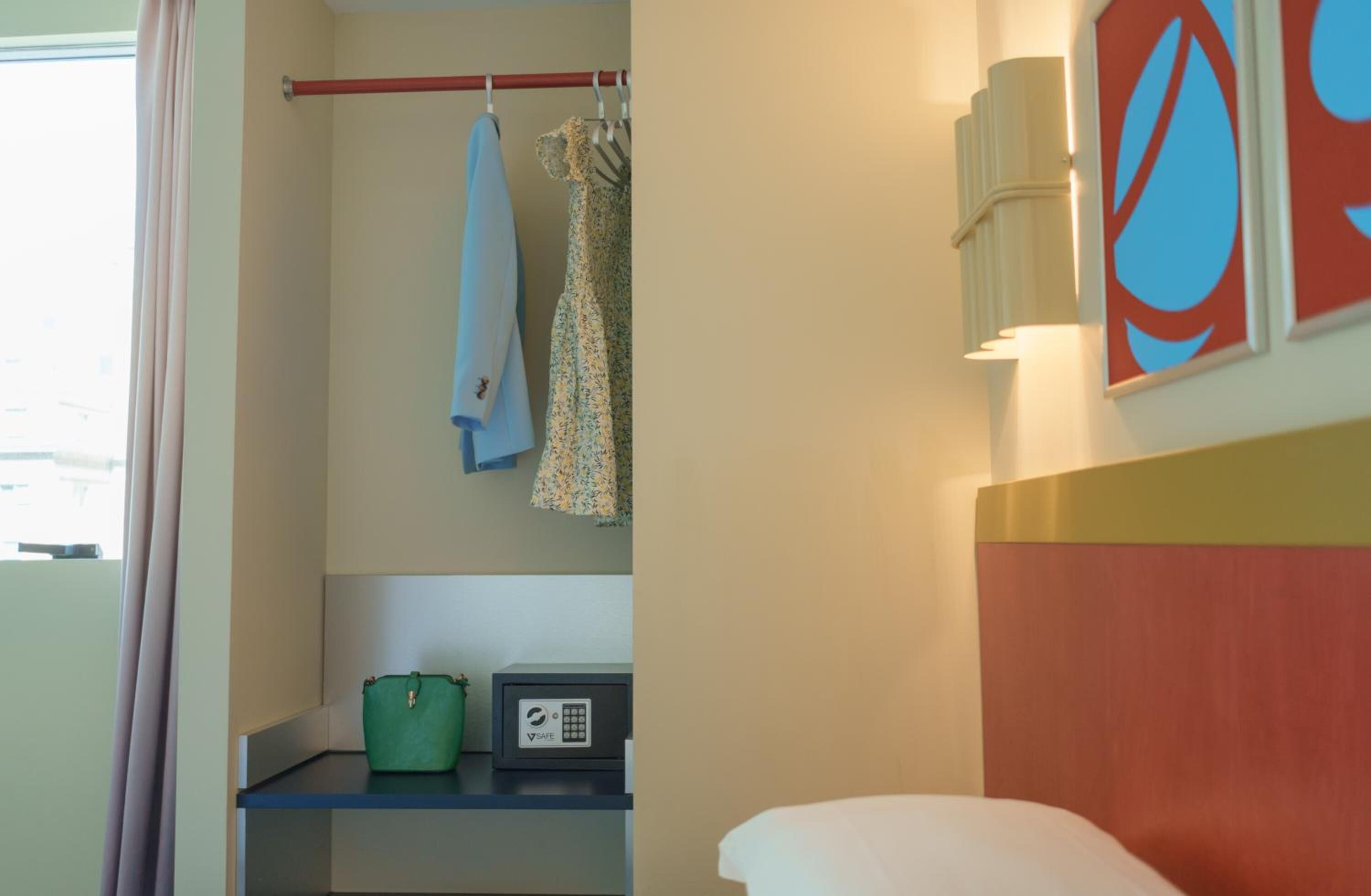 A hotel room with a bed, a small table with a green bag and a device, a coat rack with clothes, and a wall with abstract art.