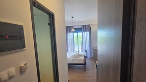 A hallway leading to a bedroom with a bed, a bedside table, and a window with curtains. There are wall-mounted light switches and a ceiling light fixture. - Falirala Central (Rhodes)
