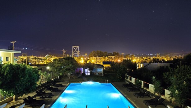 A rectangular swimming pool with a wooden deck in the foreground, surrounded by lounge chairs. In the background, there is a view of a cityscape with buildings and a bridge. - Falirala Central (Rhodes)