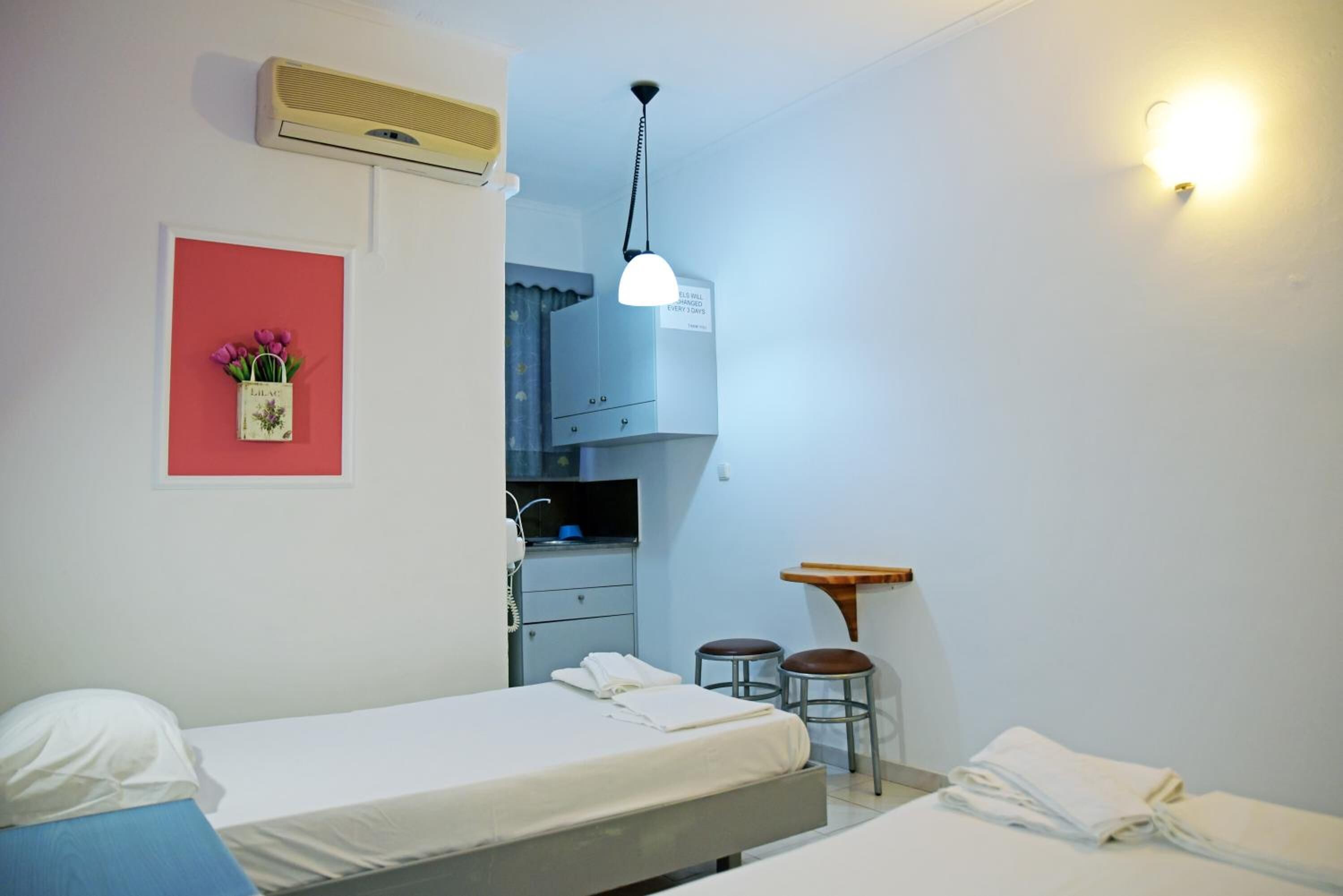 Ein Hotelzimmer mit zwei Betten, einem Schreibtisch, einem Stuhl, einem kleinen Tisch, einer Wand-Klimaanlage, einer Wandleuchte und einem Bilderrahmen mit Blumen.