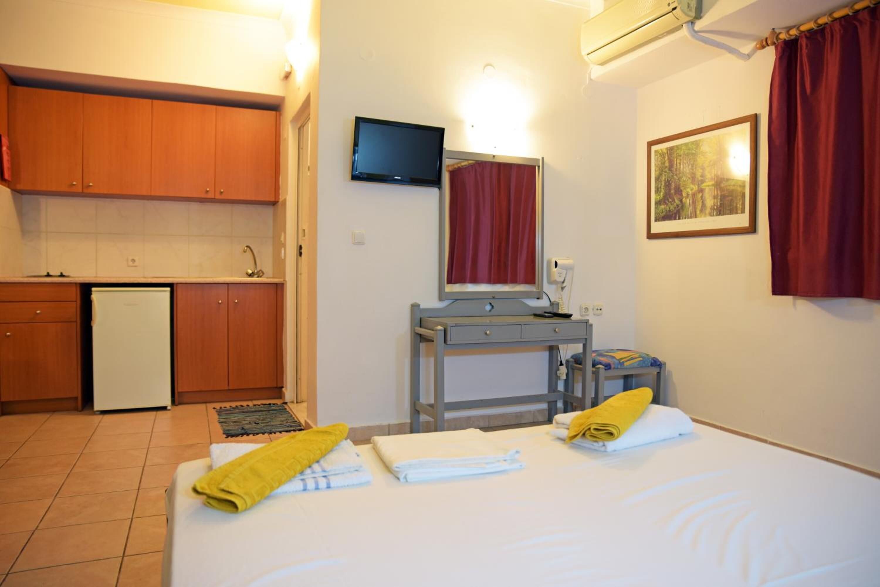 A compact hotel room with a bed, a desk, a television, a mirror, and a small kitchenette.