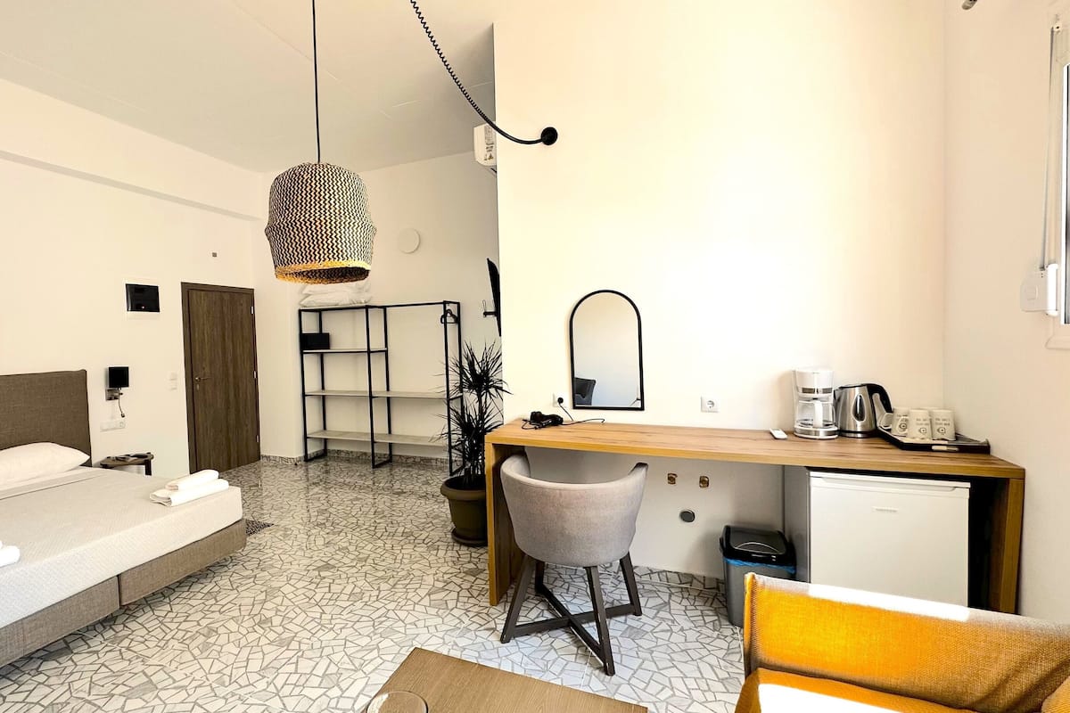 Habitación de hotel pequeña con cama, escritorio, silla, mesita, espejo, planta en maceta y cafetera.