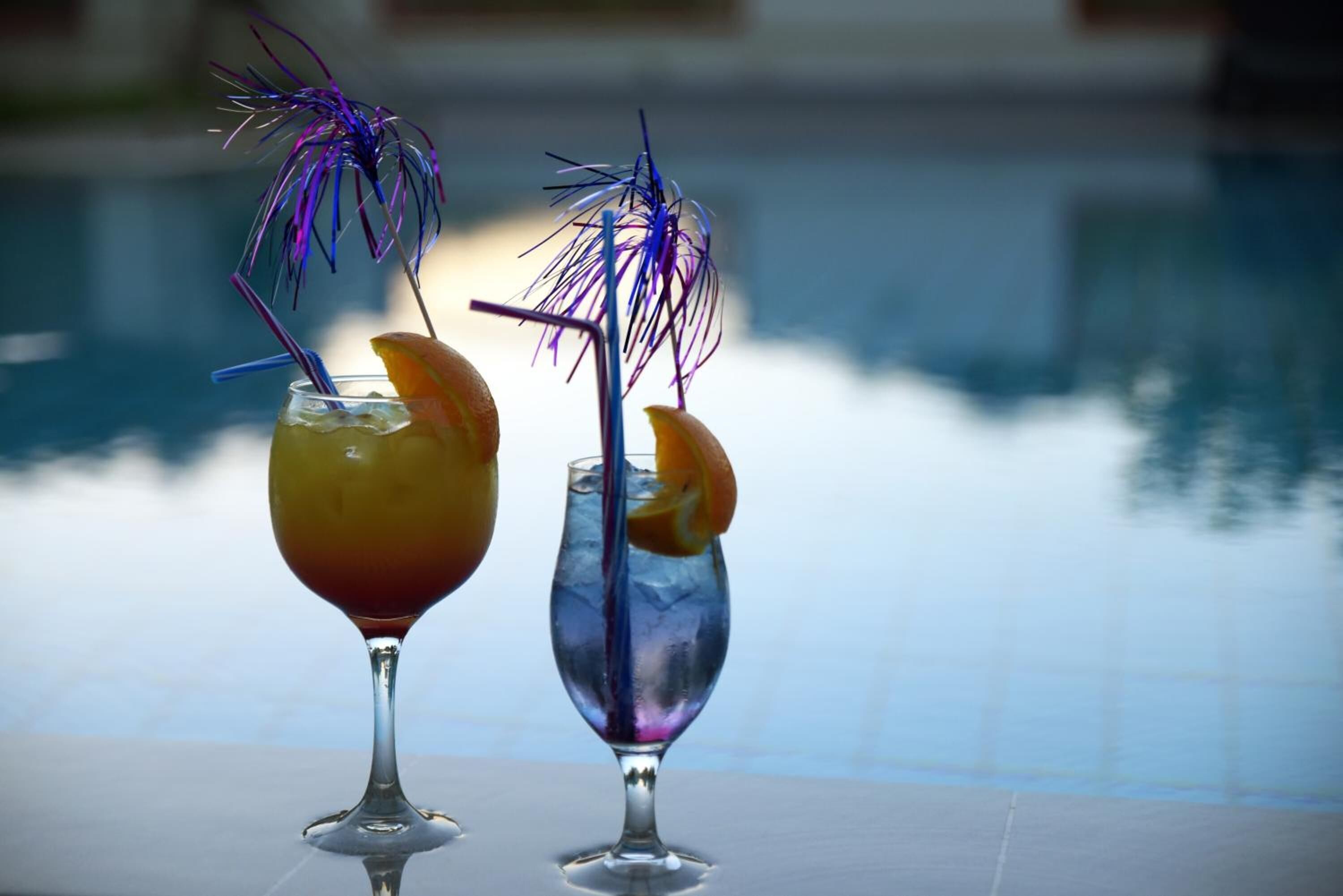 Two cocktails with straws and garnishes on a table by a pool.
