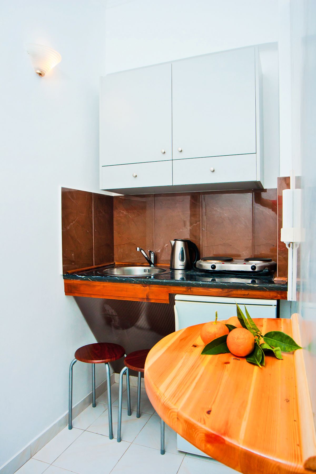A compact kitchen with a sink, stove, and cabinets. There is a wooden table with a fruit and a chair.