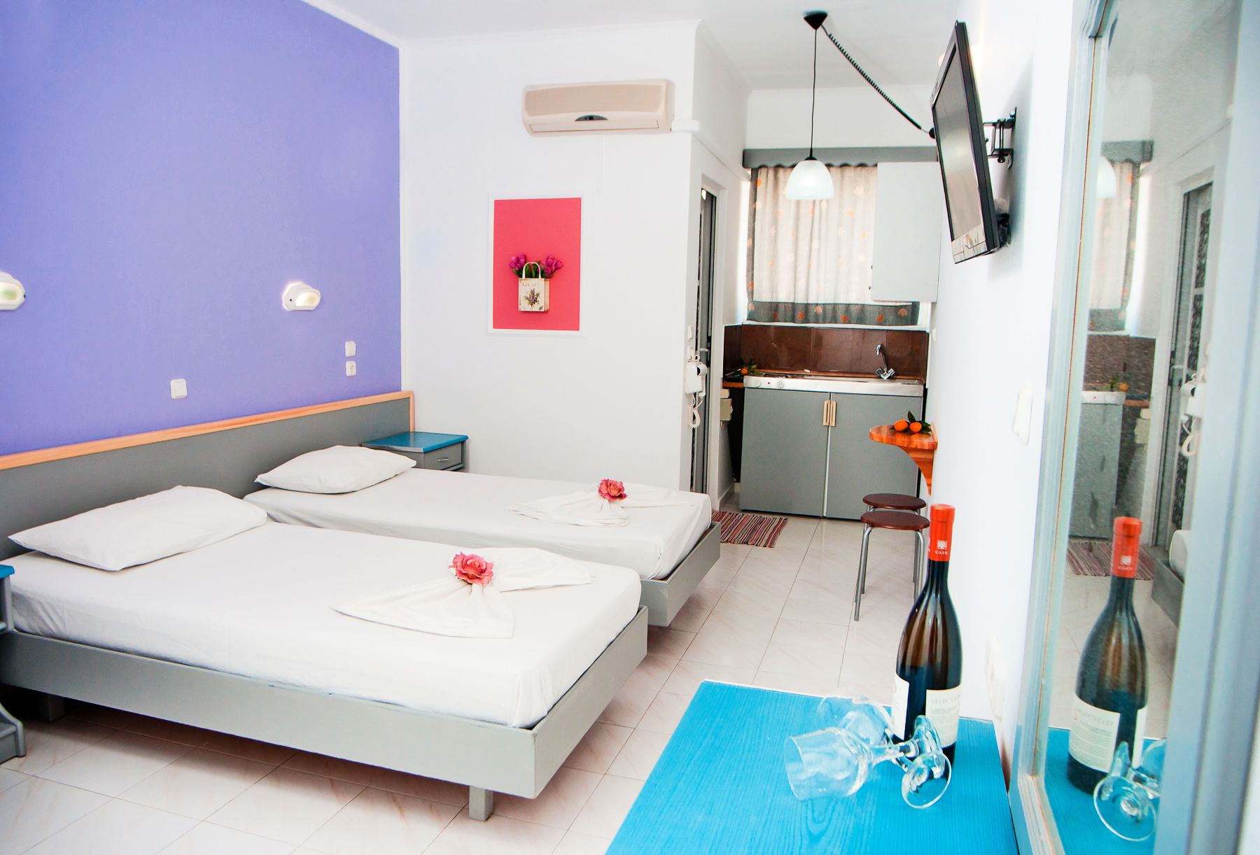 A hotel room with two beds, a kitchenette, a TV mounted on the wall, and a small table with wine bottles.