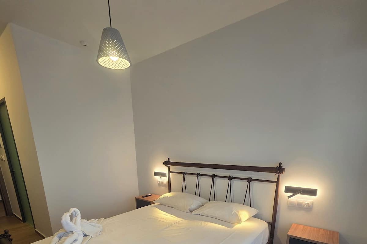 Una cama bien hecha con sábanas blancas, cabecera de madera y mesita de noche con lámpara.