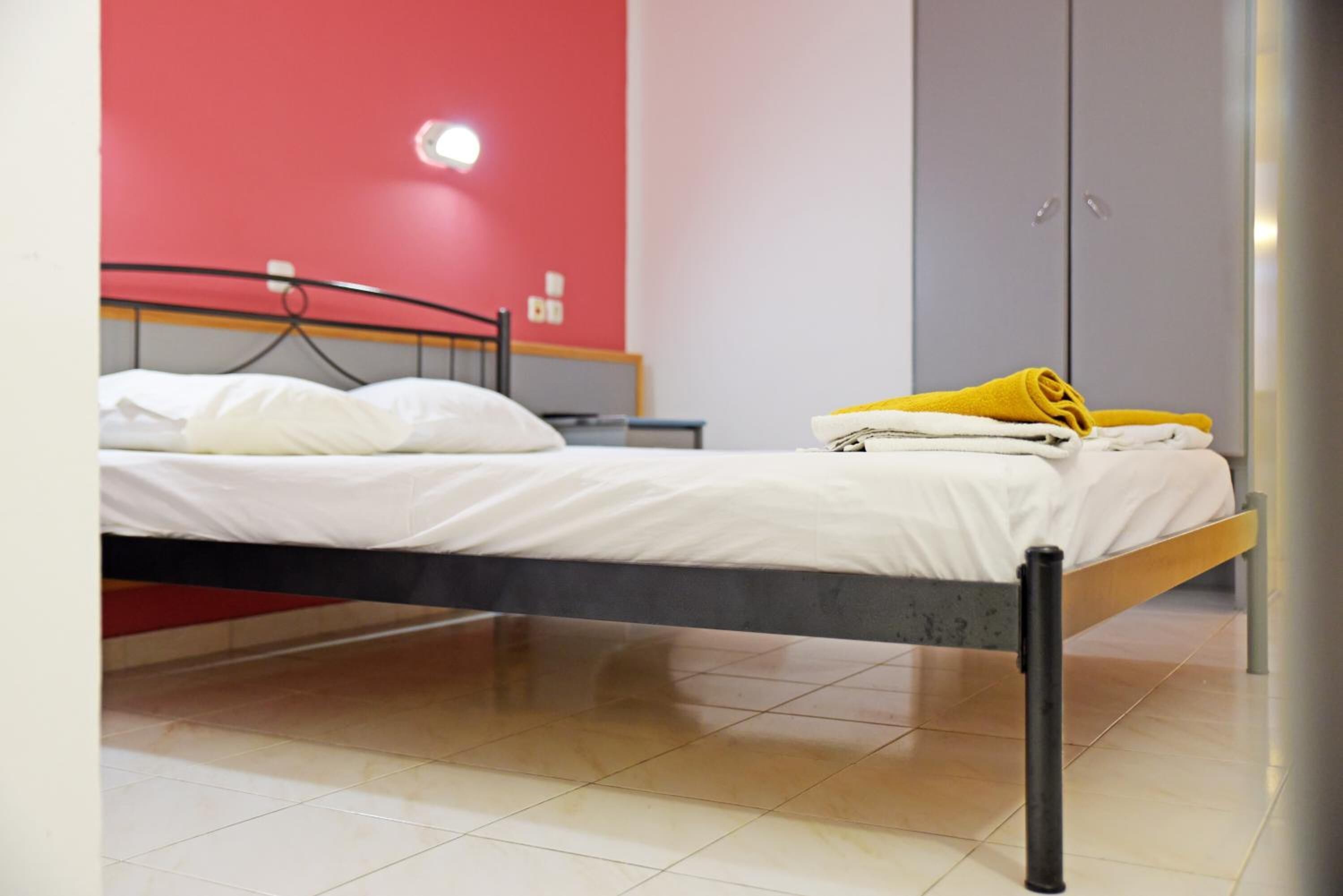 A single bed with white bedding, a yellow towel, and a red wall in the background.