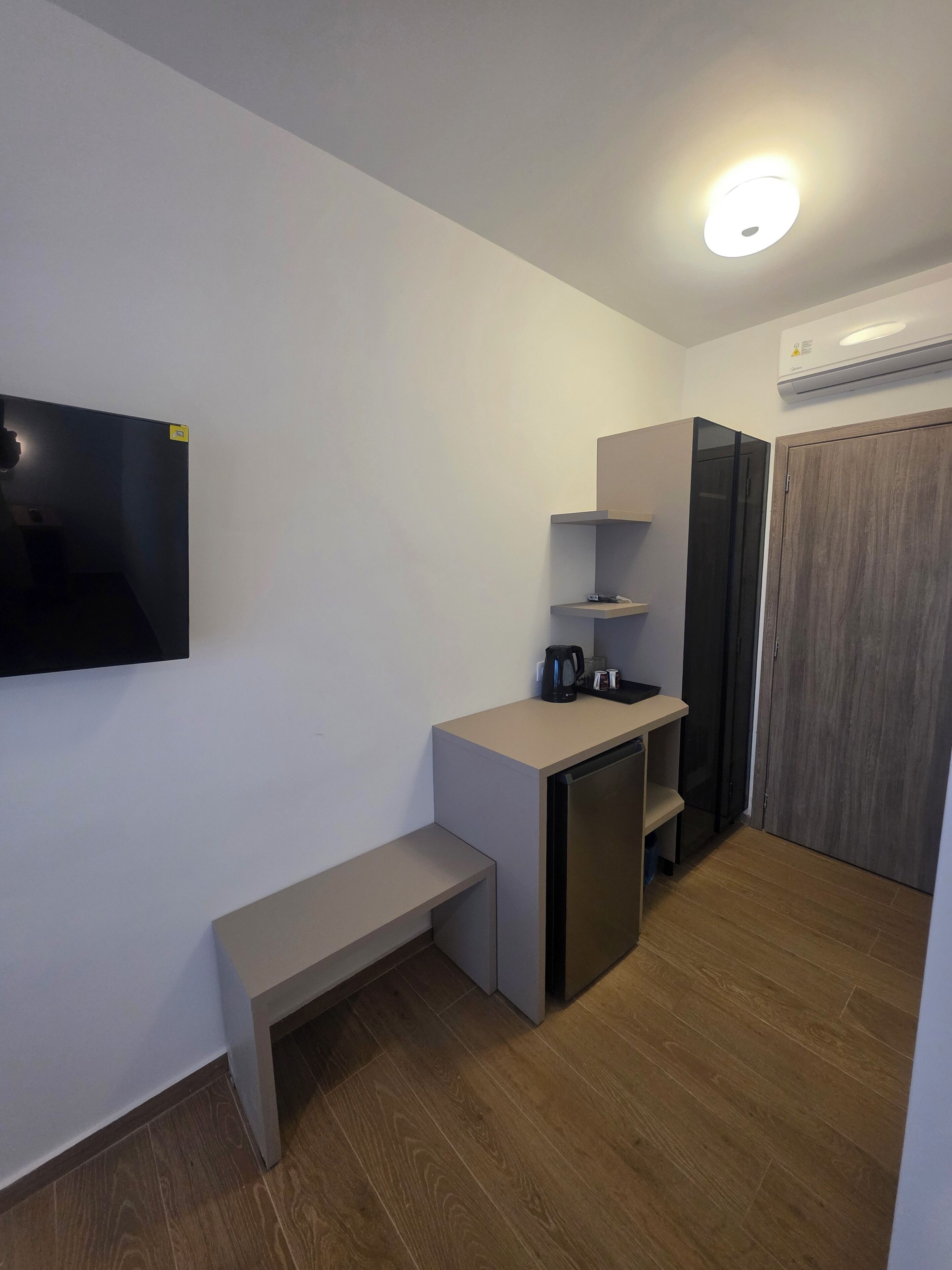 A compact kitchenette with a built-in fridge, a bench, and a flat-screen TV mounted on the wall.