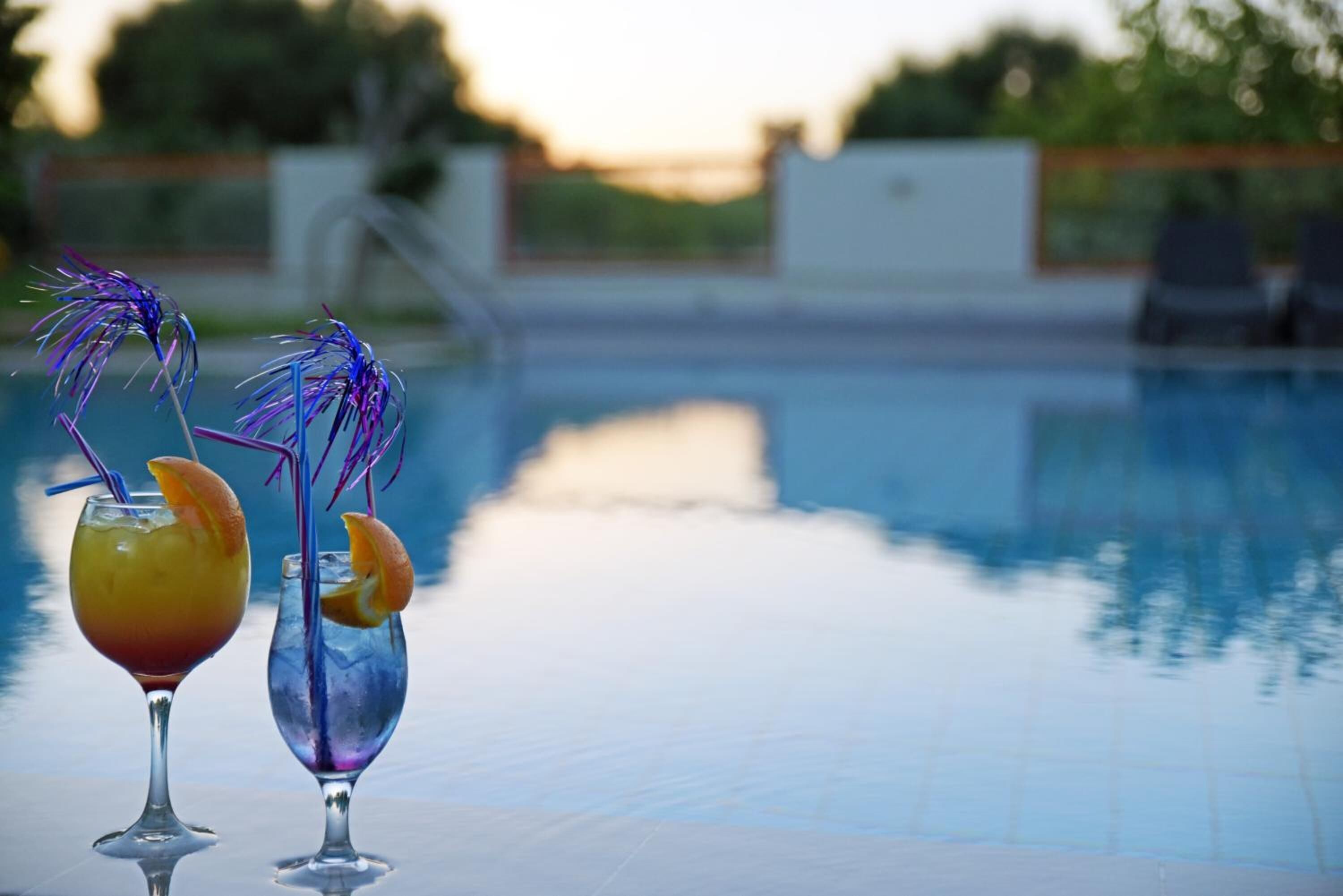 Two cocktails on a poolside table with a pool and a fence in the background.