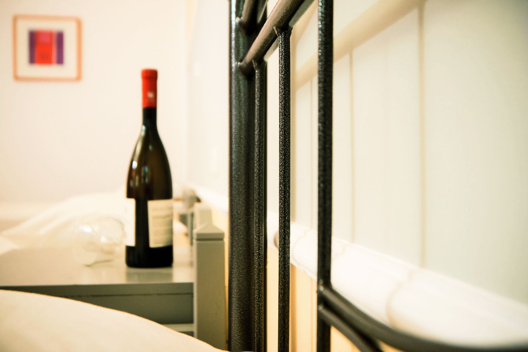 A hotel room with a bed, a bedside table with a bottle of wine, and a framed picture on the wall.